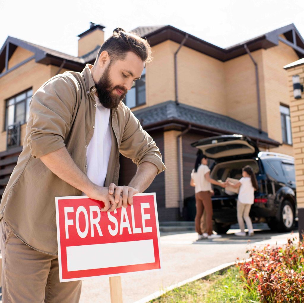 bearded man with a bun putting a for sale sign in front of a house while a woman and child load boxes into a van