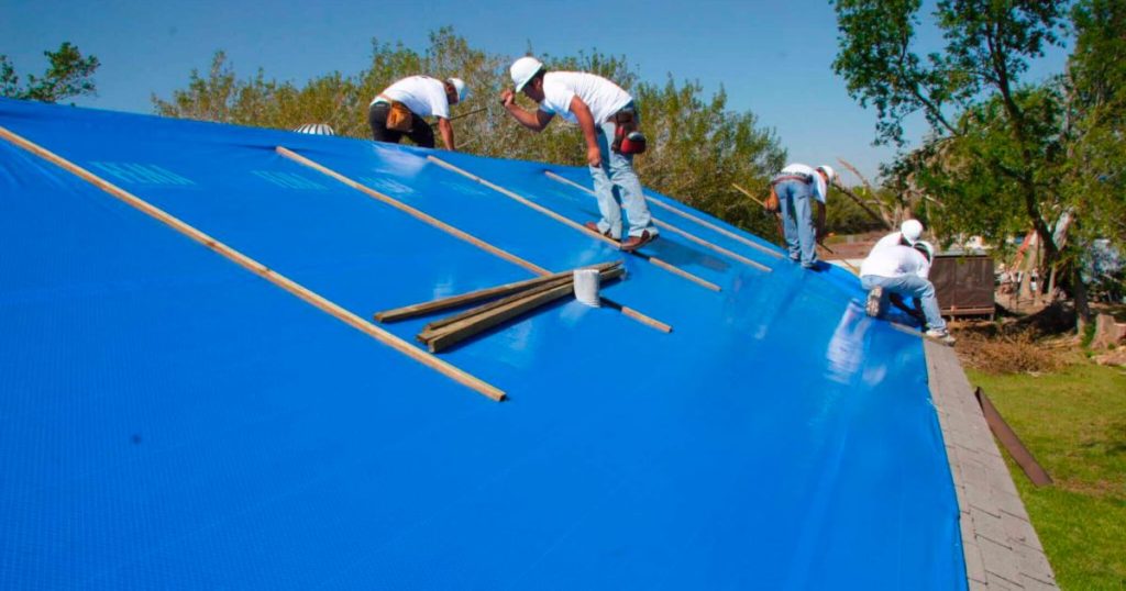 commercial roof maintenance crew of 3 roofers fastening a blue tarp to roof